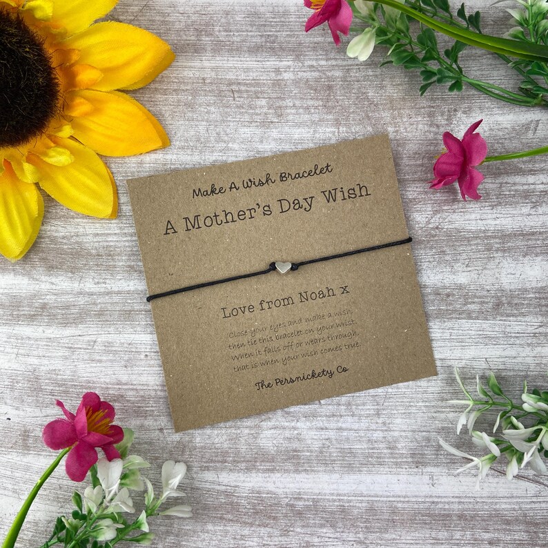 May include: A brown card with the text "Make A Wish Bracelet A Mother's Day Wish" and "Love from Noah x". The card is tied with a black string bracelet with a silver heart charm.