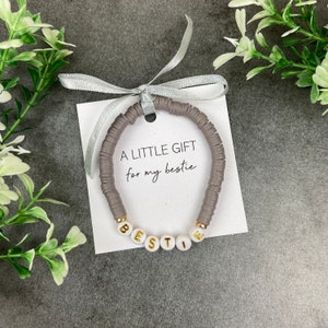 May include: A grey beaded bracelet with the word "BESTIE" spelled out in gold letters on white beads. A white card with the text "A LITTLE GIFT for my bestie" is behind the bracelet.