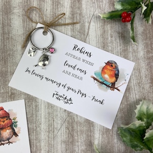 Könnte beinhalten: Eine weiße Karte mit dem Text "Rotkehlchen erscheinen, wenn geliebte Menschen in der Nähe sind" und einer Rotkehlchen-Illustration. Die Karte ist an einem silbernen Anhänger-Schlüsselanhänger mit einem Rotkehlchen-Anhänger und einem rosa Kristall befestigt. Der Text "In liebevoller Erinnerung an deinen Papa - Frank" steht unter dem Haupttext. Die Karte hat unten ein kleines Logo mit der Aufschrift "The Persnickety"