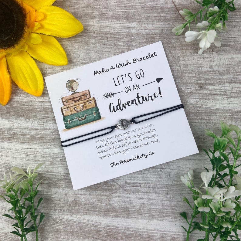 May include: A black wish bracelet with a silver charm, presented on a card with the text "Let's Go On An Adventure!". The card features illustrations of suitcases and a globe. The text on the card reads "Make A Wish Bracelet".
