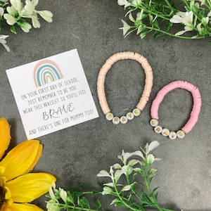 Op de afbeelding: Twee armbanden met kralen, één lichtroze en één donkerder roze, met de letters "BRAVE" in goudkleurige letters. Een kaart met een regenboogontwerp en de tekst: "OP JE EERSTE SCHOOLDAG, ONTHOUD DAT JE JEZELF MOET ZIJN. DRAAG DEZE ARMBAND ZODAT JE JE MOEDIG VOELT."