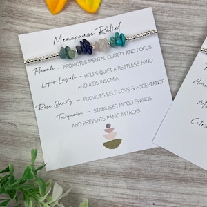 May include: A bracelet with various colored stones, including turquoise, rose quartz, lapis lazuli, and fluorite. The bracelet is accompanied by a card that lists the stones and their associated benefits for menopause relief.