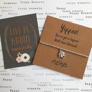 May include: A brown card with the text "Life is about moments" in gold lettering. A silver charm bracelet with a graduation cap charm is on a brown card with the text "Yippee! You've got a degree! Well done Harriet!"