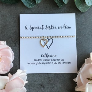 May include: A white card with the text "A Special Sister in Law" and "This little bracelet is just for you because you're my Sister in Law and I love you". A silver bracelet with two intertwined heart charms is on the card. The name "Catherine" is written on the card.