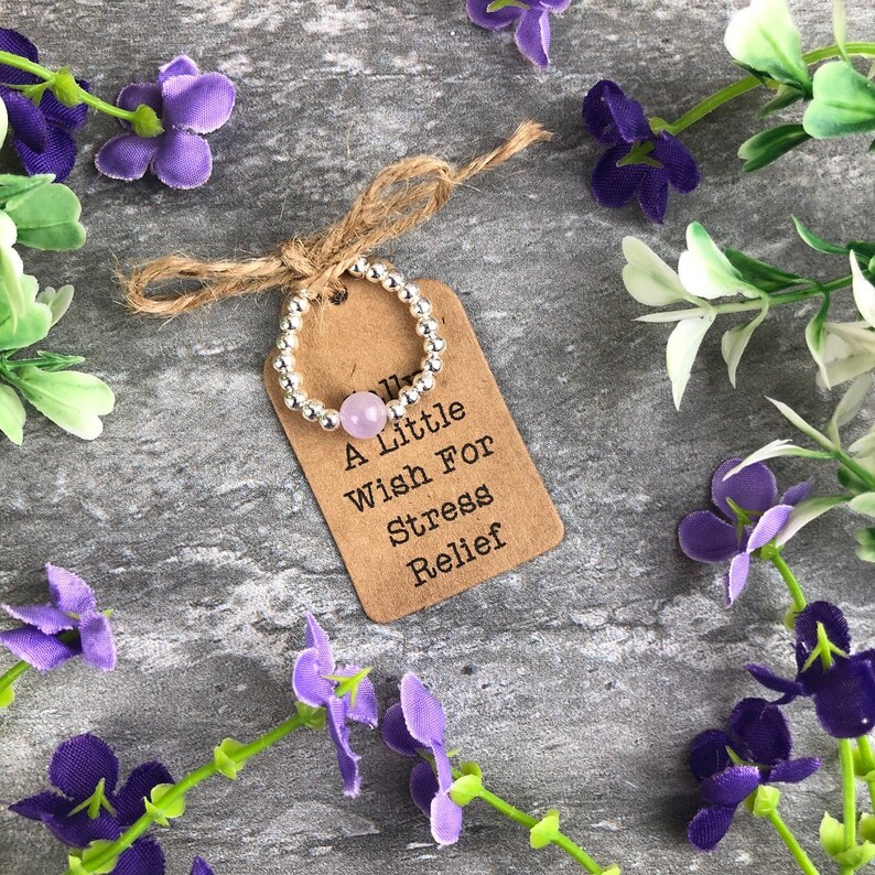 May include: A silver beaded ring with a light purple stone. The ring is on a brown tag with the text "A Little Wish For Stress Relief".