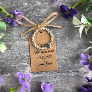 May include: A silver beaded ring with a heart charm is attached to a brown tag with the text "Auntie like you are precious and few". The tag is tied with twine and surrounded by purple and white flowers.
