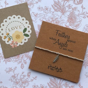 May include: A brown card with the text "Feathers appear when Angels are near" and a silver feather charm bracelet. The bracelet is on top of the card. The card has the text "The Punky Pixie Co." printed on the bottom.
