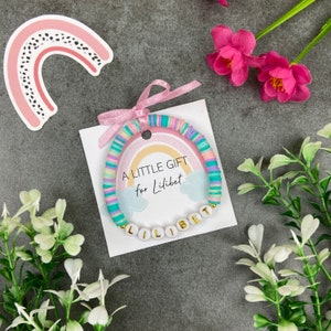 May include: A pastel rainbow bracelet with white beads spelling "Lilibet" on a pink and white card with the text "A little gift for Lilibet".