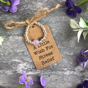 May include: A silver beaded ring with a light purple stone. The ring is tied with twine to a brown tag that reads "A Little Wish For Stress Relief".