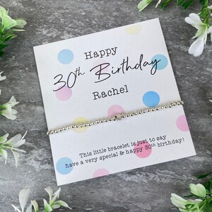 Op de afbeelding: Een zilveren kralenarmband met een sterbedel hangt op een kaart. De kaart leest "Happy 30th Birthday Rachel" met een bericht eronder. Pastelkleurige stippen versieren de kaart. De armband is een delicaat sieraad.
