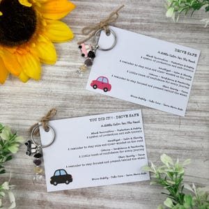 Op de afbeelding: Twee metalen sleutelhangers met kleine autobedels en edelsteenkralen bevestigd met touw aan witte kaarten. De kaarten bevatten de tekst DRIVE SAFE en YOU DID IT - DRIVE SAFE, samen met beschrijvingen van zwarte toermalijn, amethist, citrien en kwarts.