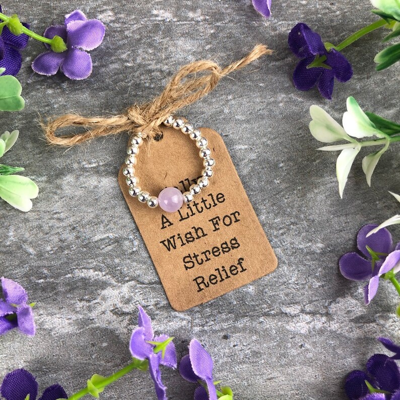 May include: A silver beaded ring with a light purple stone charm. The ring is tied with a brown twine bow and a brown paper tag that reads "A Little Wish For Stress Relief".