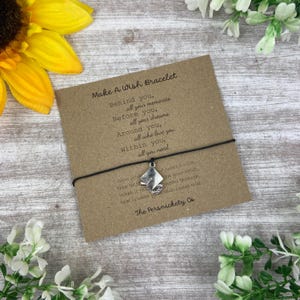 May include: A Make A Wish bracelet on a brown card with text. The black bracelet has a silver charm. The card is surrounded by a yellow sunflower and white flowers. The text on the card reads "Make A Wish Bracelet".