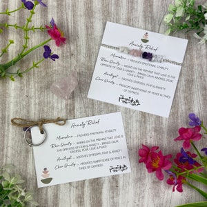 Puede incluir: Dos tarjetas blancas con el texto "Anxiety Relief" y descripciones de las propiedades de la piedra lunar, el cuarzo rosa, la amatista y el cristal de roca. Las tarjetas están al lado de un pequeño cristal de cuarzo rosa rosa y una pulsera plateada con cristales.