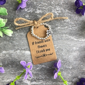 May include: A brown gift tag with the text "If friends were flowers I'd pick you" is tied with a twine bow. A silver beaded ring with a flower charm is attached to the tag. Purple flowers are scattered around.