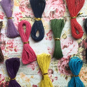 May include: Assortment of 10 spools of colorful waxed cotton cord. The cords are arranged in a semi-circle on a floral patterned surface. The colors include purple, pink, brown, dark blue, green, yellow, black, and blue.