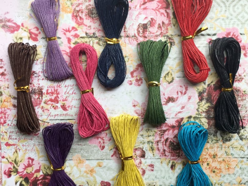 May include: Assortment of 10 spools of colorful waxed cotton cord. The cords are arranged in a semi-circle on a floral patterned surface. The colors include purple, pink, brown, dark blue, green, yellow, black, and blue.