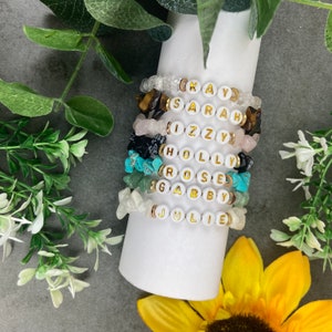 May include: A stack of seven bracelets with white beads and gold lettering spelling out the names Kay, Sarah, Izzy, Holly, Rose, Gabby, and Julie. Each bracelet is accented with different coloured gemstones.