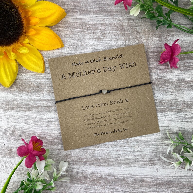 May include: A brown card with the text "Make A Wish Bracelet A Mother's Day Wish" and "Love from Noah x". A black string bracelet with a silver heart charm is attached to the card.
