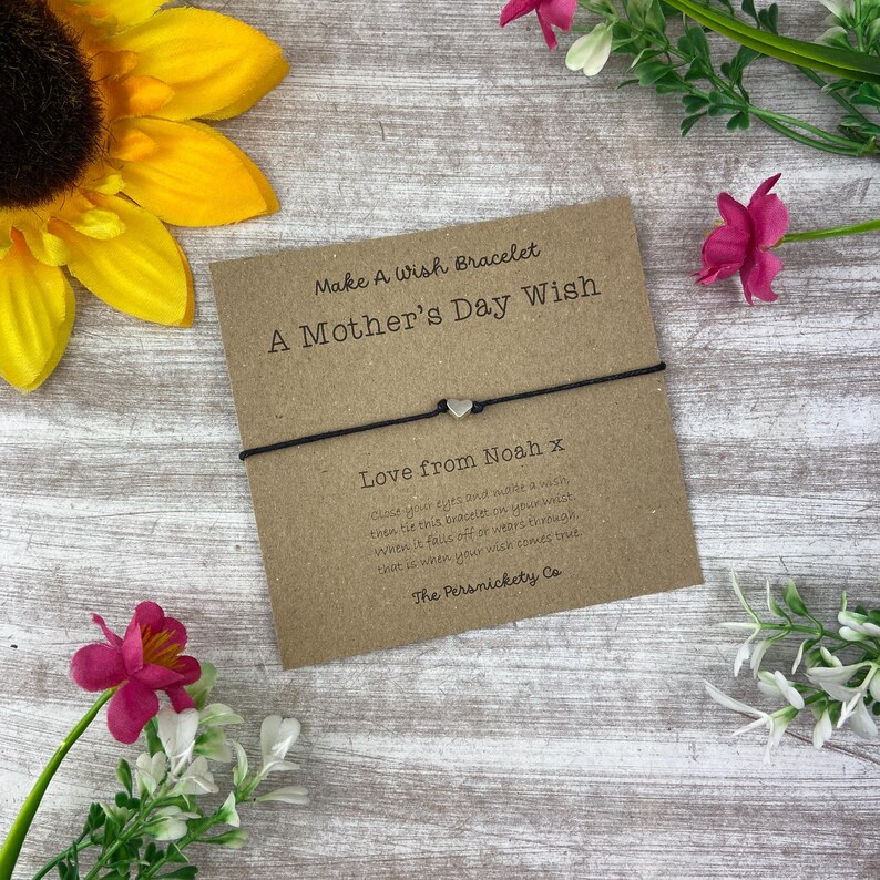 May include: A brown card with the text "Make A Wish Bracelet A Mother's Day Wish" and "Love from Noah x" with a black string bracelet with a silver heart charm.