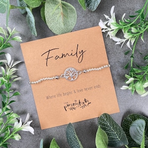 Könnte beinhalten: Ein silbernes Armband mit einem Lebensbaum-Anhänger und den Worten "Family" und "Where life begins & love never ends." Das Armband liegt auf einer braunen Karte mit dem Text "The Punsnickety Co."