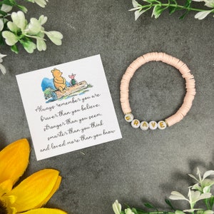 Könnte beinhalten: Ein rosa Perlenarmband mit goldenen Buchstaben, die "BRAVE" buchstabieren. Eine Karte mit einer Winnie-the-Pooh-Illustration und dem Zitat "Denke immer daran, dass du mutiger bist, als du glaubst, stärker, als du scheinst, klüger, als du denkst und mehr geliebt, als du weißt."