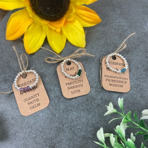 May include: Three brown cards with silver beaded rings and coloured gemstone beads. The cards have the months of February, May, and December written on them, along with the corresponding birthstones and their meanings.