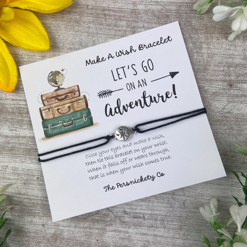 May include: A black wish bracelet with a compass charm, presented on a card with the text "Make A Wish Bracelet" and "Let's Go On An Adventure!". The card features illustrations of stacked suitcases and a globe.