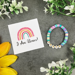 Op de afbeelding: Een witte kaart met een regenboogillustratie en de tekst "I Am Brave!" Een kleurrijke kralenarmband met het woord "Brave" gespeld in gouden letters.