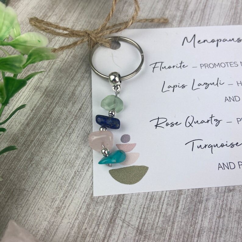 Op de afbeelding: Een zilveren sleutelhanger met een set van vijf gekleurde stenen, waaronder groen, blauw, roze en turquoise. De stenen zijn gelabeld met hun namen en eigenschappen: Fluoriet, Lapis Lazuli, Rozenkwarts en Turquoise. De kaart vermeldt ook "Menopause".