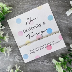May include: A white card with colorful polka dots and the text "Alice Officially a Teenager". A silver bracelet with a star charm is on the card. The text on the card reads "This little bracelet is just to say have a very special & happy 13th birthday!"