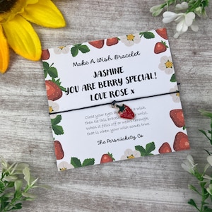 May include: A white card with a strawberry and floral design, featuring the text "Make A Wish Bracelet" and "JASMINE YOU ARE BERRY SPECIAL! LOVE ROSE x". A black string bracelet with a red strawberry charm is attached.