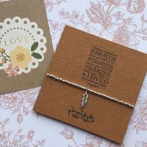 May include: A silver bracelet with a small feather charm. The bracelet is on a brown card with the text "Always remember you are BRAVER than you BELIEVE STRONGER than you SEEM SMARTER than you THINK & LOVED more than you know" and the logo "The Persnickety Pixie".