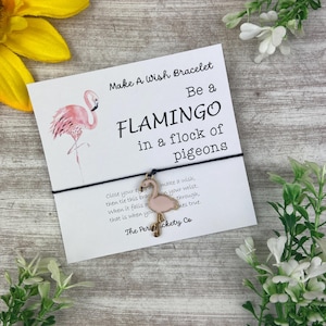 Op de afbeelding: Roze flamingo wensarmband met een zwart koord en een kaart met de tekst "Make A Wish Bracelet Be a Flamingo in a flock of pigeons" en "Close your eye make a wish, then tie this bracelet on your wrist. When it falls off through, that is when your wish comes true. The Perickety Co."