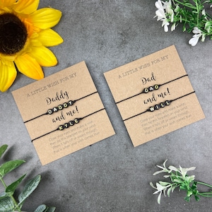 以下が含まれることがあります： 茶色のカードに「Daddy and me!」と「Kyle」と「Dad and me!」と「Will」と書かれた文字ビーズが付いた2つの黒いストリングブレスレット。「A Little Wish For My」と書かれたカード。背景にはヒマワリと白い花が含まれています。