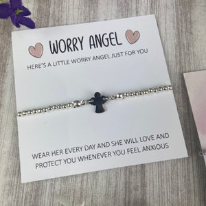 May include: A white card with the text "Worry Angel" and two pink hearts. The card is next to a silver beaded bracelet with a small silver angel charm. The text on the card reads "Here's a little worry angel just for you. Wear her every day and she will love and protect you whenever you feel anxious."