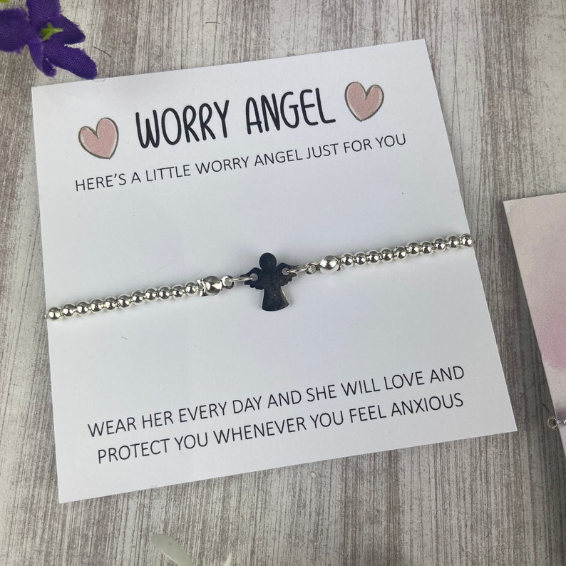 May include: A white card with the text "Worry Angel" and two pink hearts. The card is next to a silver beaded bracelet with a small silver angel charm. The text on the card reads "Here's a little worry angel just for you. Wear her every day and she will love and protect you whenever you feel anxious."