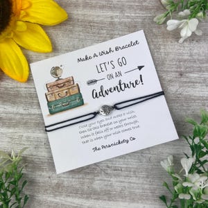 May include: A black wish bracelet with a silver compass charm, presented on a card with the text "Let's Go On An Adventure!". The card features illustrations of stacked suitcases and a globe, with instructions for use.