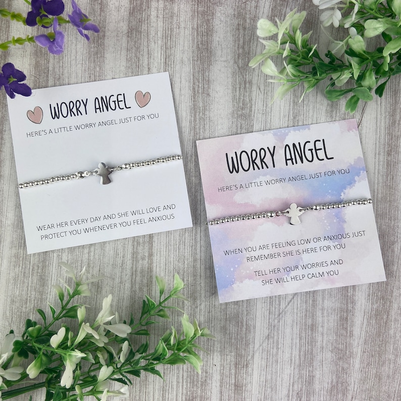 May include: Two silver bracelets with angel charms and inspirational messages. The bracelets are on white cards with pink hearts and a watercolour background. The text on the cards reads "Worry Angel" and "Here's a little worry angel just for you".