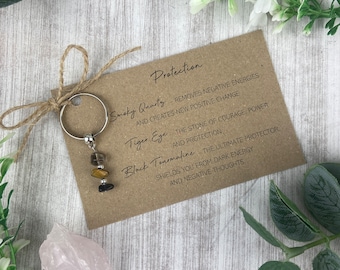 May include: A brown card with the text "Protection" and a list of crystals and their properties. The crystals are smoky quartz, tiger eye, and black tourmaline. The card is next to a silver key chain with the same crystals attached.