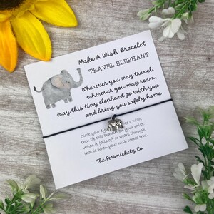 May include: A white card with black text that reads "Make A Wish Bracelet Travel Elephant" and "Wherever you may travel, wherever you may roam, may this tiny elephant go with you and bring you safely home."  The card has a small silver elephant charm attached to a black string.