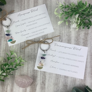 Op de afbeelding: Twee sleutelhangers met verschillende gekleurde stenen en metalen ringen. De sleutelhangers zijn gelabeld met de woorden "Menopause Relief" en "Perimenopause Relief". Elke sleutelhanger heeft een lijst met stenen en hun bijbehorende voordelen.