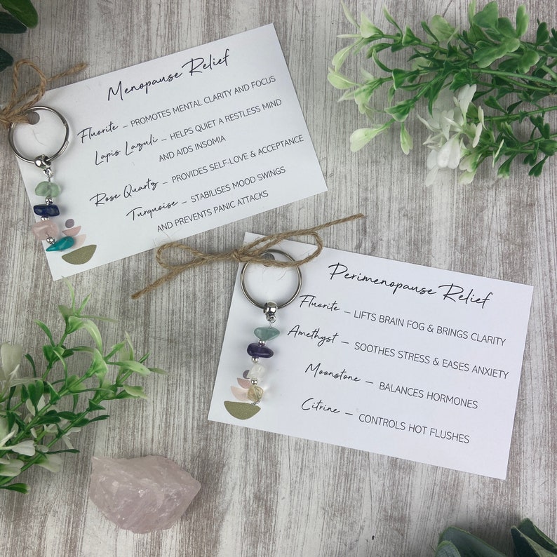 Op de afbeelding: Twee sleutelhangers met verschillende gekleurde stenen en metalen ringen. De sleutelhangers zijn gelabeld met de woorden "Menopause Relief" en "Perimenopause Relief". Elke sleutelhanger heeft een lijst met stenen en hun bijbehorende voordelen.