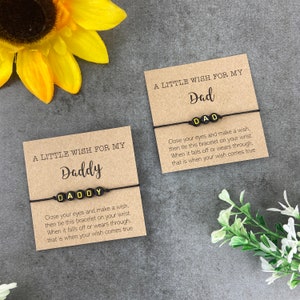 Könnte beinhalten: Zwei Karten mit dem Text "A Little Wish For My" und entweder "Daddy" oder "Dad" darauf. Jede Karte hat ein schwarzes Armband aus Schnur mit gelben Buchstabenperlen, die das Wort auf der Karte buchstabieren.