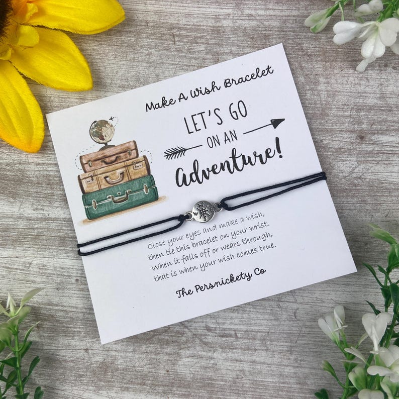 May include: A black cord wish bracelet with a silver-colored charm, presented on a white card. The card features the text "Make A Wish Bracelet" and "Let's Go On An Adventure!" with illustrations of luggage and a globe. The Persnickety Co.