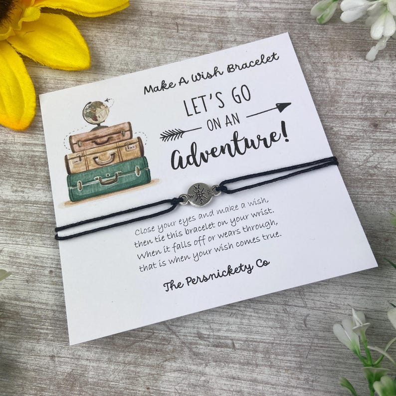 May include: A black wish bracelet with a silver compass charm, presented on a card with the text "Make A Wish Bracelet" and "Let's Go On An Adventure!". The card features illustrations of stacked vintage suitcases.