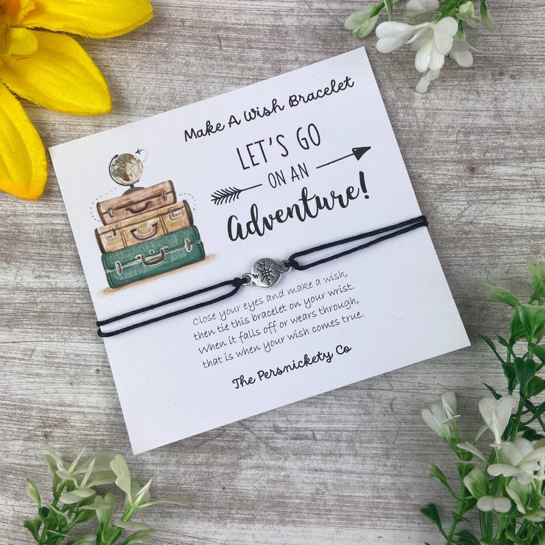 May include: A black wish bracelet with a silver charm, displayed on a white card. The card features the text "Make A Wish Bracelet" and "LET'S GO ON AN Adventure!" with a graphic of stacked suitcases. The Persnickety Co.