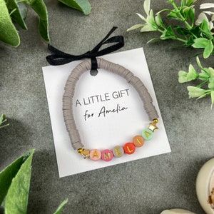 Peut inclure: Un bracelet de perles grises avec le nom "Amelia" épelé avec des perles colorées et des lettres dorées. Le bracelet est attaché à une carte blanche avec un ruban noir et le texte "A LITTLE GIFT for Amelia".