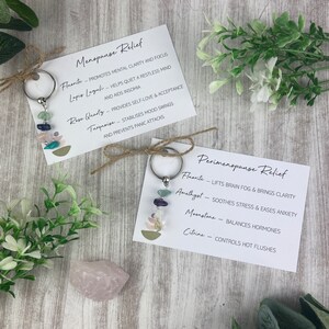 Op de afbeelding: Twee sleutelhangers met verschillende sets kristallen en beschrijvingen van hun voordelen voor het verlichten van menopauze en perimenopauze. De sleutelhangers zijn gemaakt met zilverkleurige metalen ringen en bevatten verschillende gekleurde kristallen, waaronder blauw, groen, paars en wit.