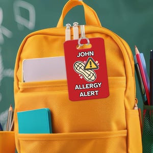 Può includere: Uno zaino giallo con un'etichetta rossa di allerta allergia che dice "JOHN ALLERGIA ALERT" con un'immagine di arachidi e un punto esclamativo. L'etichetta è attaccata allo zaino con un anello di plastica trasparente.